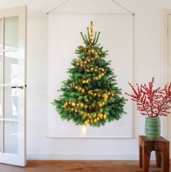Levensechte kerstboom, gedrukt op stof. Met lichtjes erin. Deze hangt aan de muur en ervoor staat een groene vaas met rode bessentakken.