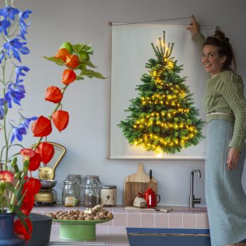 Kerstboom poster met lichtjes wordt opgehangen door vrouw in kleurrijke keuken.