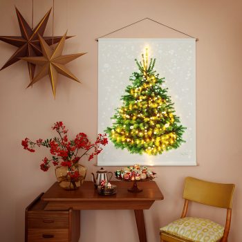 Kerstboom wandkleed met kerstverlichting hangt op een roze muur. Links 2 papieren kerststerren. Op het vintage bureautje staat een schaal met mooie oude kerstballen.