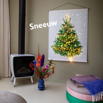 Alternatieve kerstboom (wandkleed) met sneeuwvlokken eroverheen gedrukt. Dit wandkleed (met lichtjes!) hangt op een groene muur. Ervoor staat een gekleurde poef en een fleurige bos bloemen, en een kacheltje. Winters binnen - gezellig.