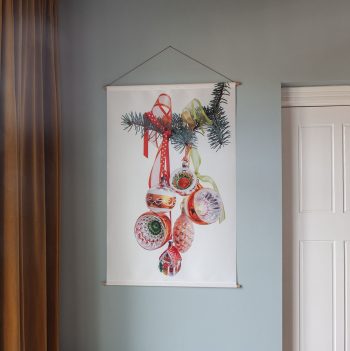 Er hangt een wandkleed op een lichtblauwe muur. Op het wandkleed staat een foto van een tros vintage kerstballen (hangend aan een kersttakje)