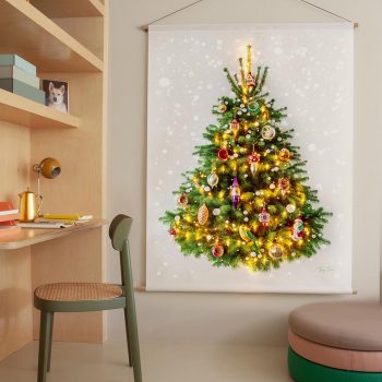 Kerstboom op doek - met kerstballen en kerstlichtjes, supergezellig. Dit kerstboomwandkleed hangt aan de muur. Rechts een kast met stoel (een thuiswerkplek) en links een gekleurde poef.