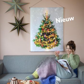 Gezellig kerstboom-wandkleed boven een bank. Waar een vrouw, onder een warm dekentje, een boek leest met ee kop thee in haar handen. De muur is roze, er hangen ook 2 kerststerren, heel gezellig. De kerstboom zit vol prachtige ouderwetse kerstballen en lichtjes. Deze zijn OP het doek gedrukt. Net echt. De vrouw kijkt vrolijk in de camera.