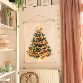 Er hangt een heel gezellig kerstboomwandkleed aan de muur. Deze is versierd met prachtige oude kerstballen en lichtjes. (Deze zijn op het doek gedrukt, net echt!) Ook dwarrelen er subtiele sneeuwvlokken over het doek Heel gezellig. Links staat een vitrinekast met mooi oud serviesgoed.