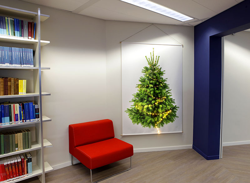 Duurzame Kerstversiering kantoor Duurzame kerstboom op kantoor. Met links een grote boekenkast en een rood bankje. Rechts een blauwe balk. En in het midden de Tiny-Trees kerstboom-wandkleed.